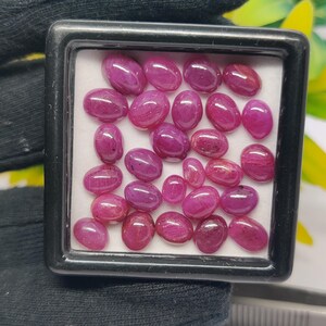 May include: A collection of oval-shaped, polished ruby gemstones in varying sizes. The rubies are a deep, rich red color and are displayed in a black, square display case with a white interior.