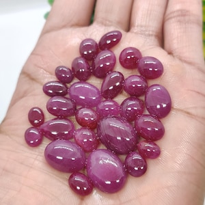 Könnte beinhalten: Eine Sammlung ovaler und tropfenförmiger, polierter Rubin-Edelsteine in verschiedenen Größen. Die Edelsteine haben eine tiefe, satte rote Farbe mit glänzendem Finish, in einer Hand präsentiert.