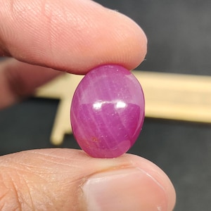 以下が含まれることがあります： 磨かれた楕円形のルビーの宝石。宝石は、滑らかで反射する表面を持つ、深く鮮やかなピンクがかった赤色です。ルビーは2本の指の間に挟まれ、そのサイズと形状を示しています。