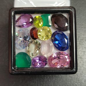 Può includere: Una collezione di varie pietre preziose in una vetrina nera. Le pietre preziose sono di diverse forme e colori, tra cui viola, verde, giallo, blu e rosa. Le pietre preziose sono disposte su uno sfondo bianco.