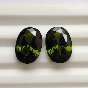 Puede incluir: Dos gemas de corte ovalado con un tono verde oscuro y superficies facetadas. Las gemas tienen un interior verde lima vibrante, sobre un exterior profundo, casi negro. Las piedras se exhiben sobre una superficie blanca.
