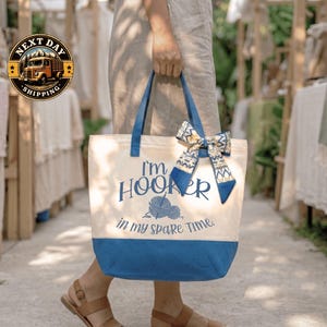 A person is holding a blue and white tote bag with the text "I'm Hooker in my spare time" printed on it.