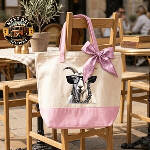 A pink tote bag with a cartoon goat wearing sunglasses and a bow tie, sitting on a wooden chair.
