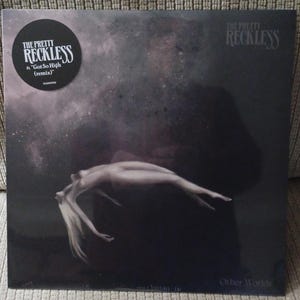 May include: Album cover for "Other Worlds" by The Pretty Reckless. The cover features a grayscale image of a person floating in a dark, starry space. A circular sticker on the top left corner displays the band's name and the remix title.