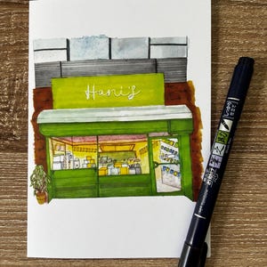 May include: Watercolor illustration of a storefront with a green awning and the name "Hani's" in white. A black pen with Japanese text is on the right side of the image. The storefront has a brown brick facade and a small potted plant.