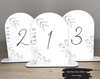 May include: Three white table number signs with black floral designs and brown numbers. The numbers are "1", "2", and "3". The text "TABLE" is written vertically on each sign.
