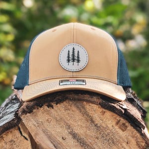 May include: A tan and navy blue trucker hat with a circular patch featuring a tree design. The hat is resting on a pile of cut logs, with a blurred green background.