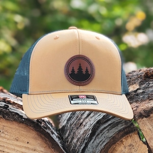 May include: A tan and navy blue trucker hat with a circular patch featuring a forest of trees. The hat has a curved brim and a mesh back. The patch is brown with a black tree design. The hat is sitting on a pile of wood.