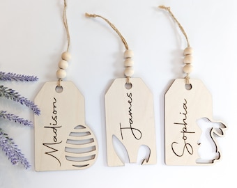 May include: Three wooden Easter tags with names "Madison", "James", and "Sophia" written on them. Each tag has a different design cut out: an Easter egg, a bunny, and a carrot. The tags are strung with twine and wooden beads.