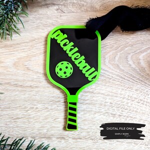 Puede incluir: Adorno de raqueta de pickleball verde y negro con la palabra "pickleball" escrita en diagonal sobre la cara de la raqueta. Una pelota de pickleball verde est&aacute; en el centro de la raqueta.