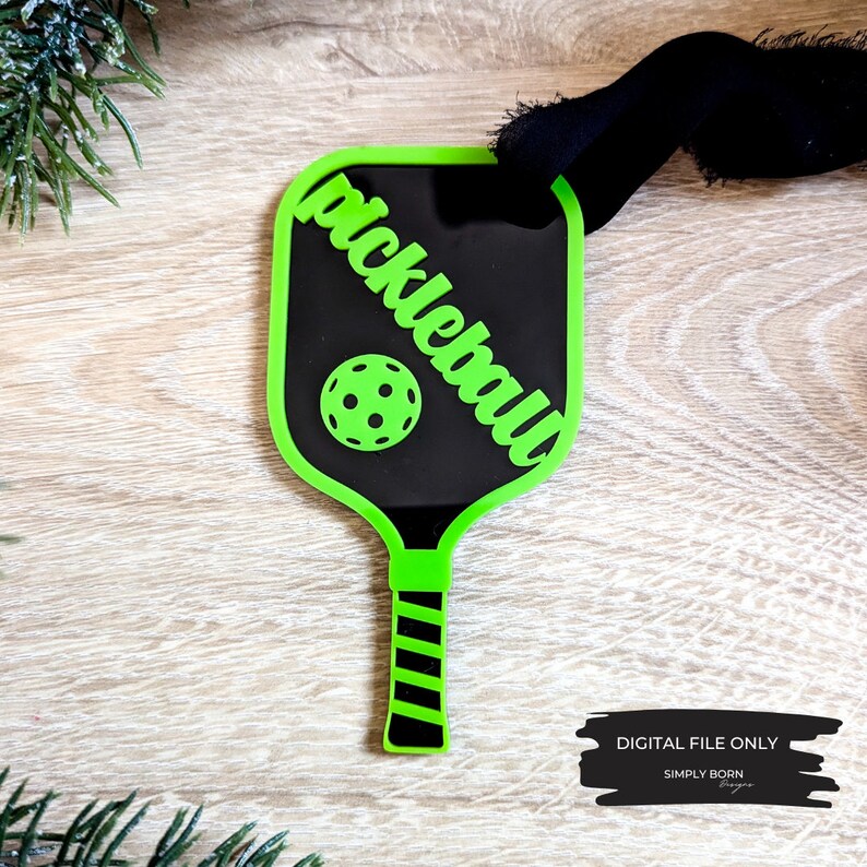Puede incluir: Adorno de raqueta de pickleball verde y negro con la palabra "pickleball" escrita en diagonal sobre la cara de la raqueta. Una pelota de pickleball verde est&aacute; en el centro de la raqueta.