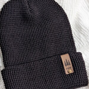 May include: A black knit beanie with a textured waffle pattern and a folded cuff. A rectangular tan leather patch is attached to the cuff, featuring an embossed graphic of three pine trees above the letters PNW and a small metallic rivet.