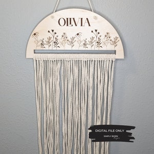 May include: A wooden half-circle wall hanging with the name "Olivia" engraved on it. The hanging features a row of small flowers and leaves along the top edge and a fringe of white yarn hanging from the bottom.