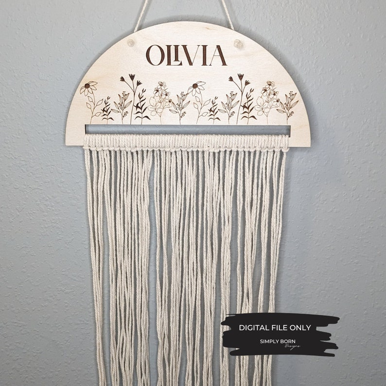 May include: A wooden half-circle wall hanging with the name "Olivia" engraved on it. The hanging features a row of small flowers and leaves along the top edge and a fringe of white yarn hanging from the bottom.