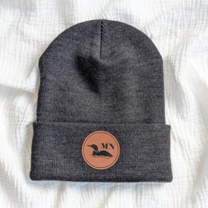 May include: A charcoal gray knit beanie with a folded cuff. Centered on the cuff is a circular tan leather patch featuring a black silhouette of a loon swimming on water and the letters MN printed in a bold, dark font above the bird.