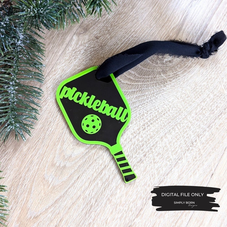 Puede incluir: Adorno de raqueta de pickleball negro y verde con una cinta negra. La raqueta tiene la palabra "pickleball" escrita en ella en letras verdes.