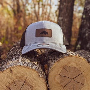 May include: A gray and black trucker hat with a brown leather patch featuring a black silhouette of a state outline. The hat is sitting on top of two stacked logs.