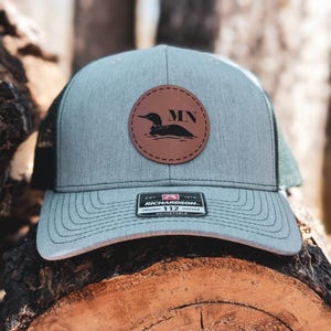 May include: A gray trucker hat with a brown leather patch featuring a black loon and the letters "MN". The hat has a curved brim and a mesh back. The hat is adjustable.