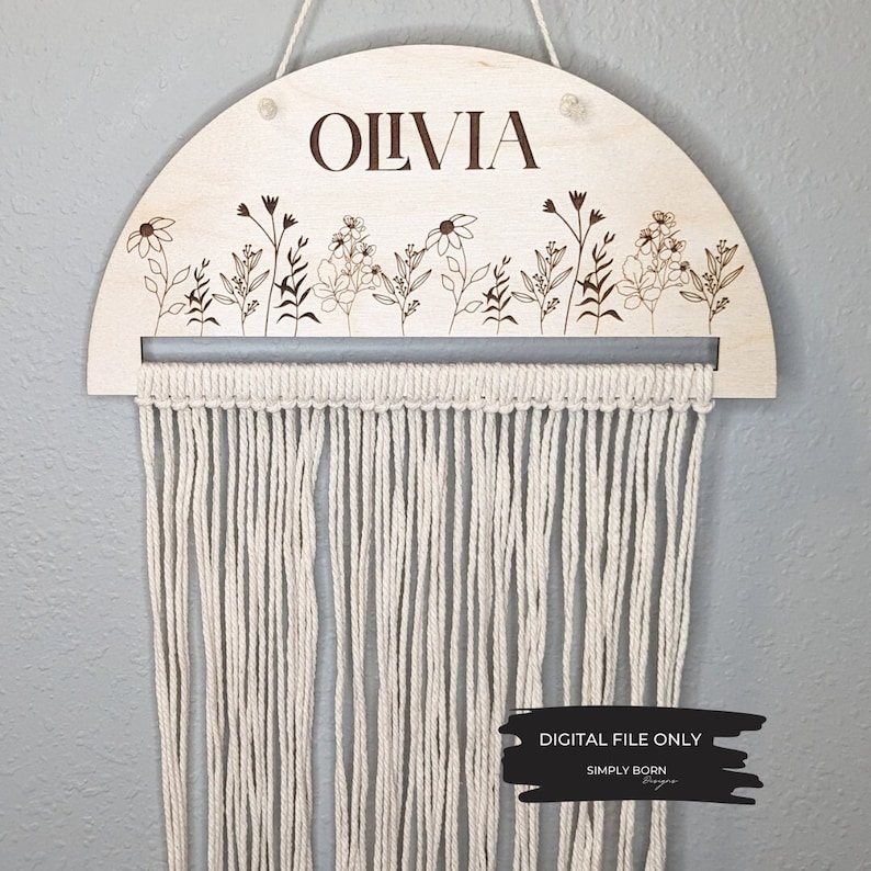 May include: A wooden half-circle wall hanging with the name "Olivia" engraved on it. The hanging features a floral design and a macrame fringe.