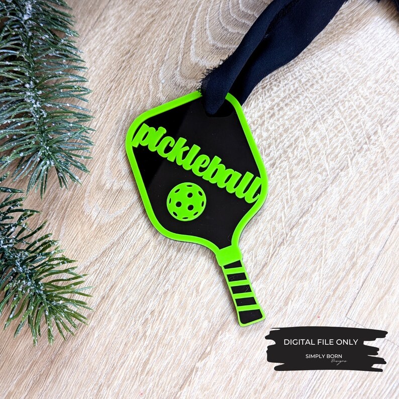 Puede incluir: Un adorno de raqueta de pickleball verde y negro con la palabra "pickleball" escrita en verde en la raqueta. La raqueta tiene un mango rayado en blanco y negro.