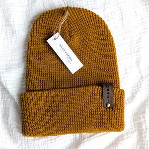 May include: A mustard yellow waffle knit beanie with a brown leather tag that says "56431". The beanie has a white tag attached to it that says "SIMPLY BORN Designs."