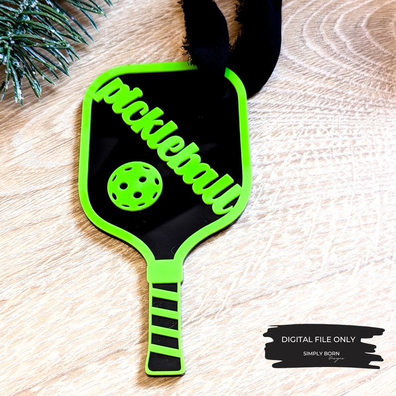 Puede incluir: Adorno de raqueta de pickleball verde y negro con una cinta negra. La raqueta tiene la palabra "pickleball" escrita en ella en una fuente verde cursiva.