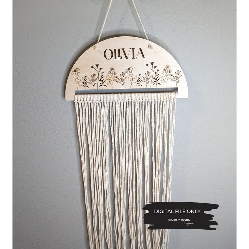 May include: A wooden half-circle wall hanging with the name "Olivia" engraved on it. The hanging features a floral design and a fringe of white yarn.