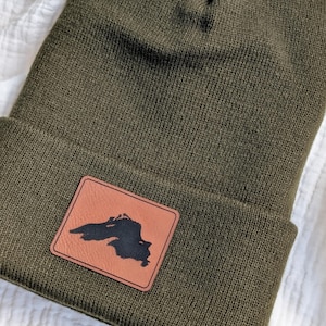 May include: An olive green knit beanie with a folded cuff. Centered on the cuff is a rectangular tan leather patch featuring a black silhouette graphic of Lake Superior. The beanie has a ribbed texture and a classic cuffed design.