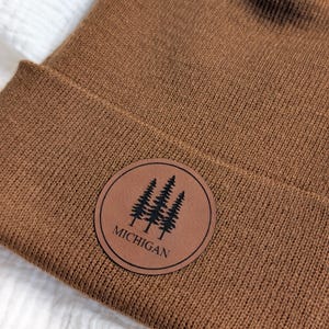 May include: A brown knit beanie featuring a circular brown patch on the cuff. The patch displays a black graphic of three pine trees above the text MICHIGAN in a simple sans-serif font. The beanie has a ribbed texture and a folded cuff design.