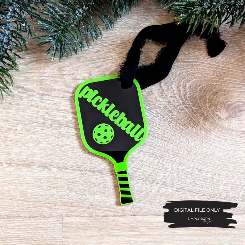 Puede incluir: Adorno de raqueta de pickleball verde con fondo negro y la palabra "pickleball" escrita en blanco. La raqueta tiene un mango rayado en blanco y negro y una pelota de pickleball negra en el centro.