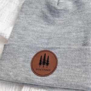 May include: A light grey knit beanie featuring a circular brown faux leather patch centered on the cuff. The patch displays a black graphic of three pine trees above the text WISCONSIN, all enclosed within a thin black circular border line.