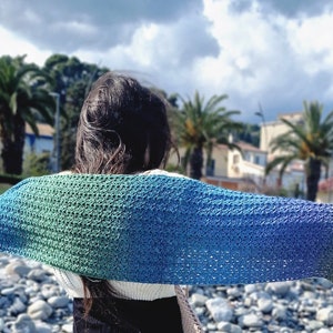 May include: A woman wearing a colourful crochet shawl with a gradient of green, blue and yellow. The shawl is draped over her shoulders and she is standing on a beach with rocks in the foreground.