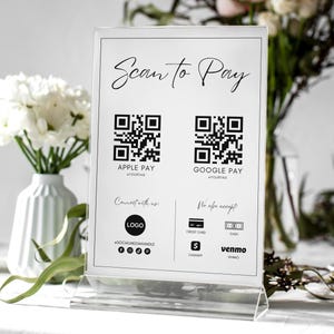 May include: White sign with black text that reads "Scan to Pay". Two black and white QR codes are displayed with the text "Apple Pay" and "Google Pay" below each. The sign also lists other payment methods accepted, including credit card, cash, Venmo, and a logo for a social media handle.