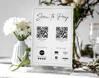 Op de afbeelding: Een wit bord met zwarte tekst die "Scan to Pay" luidt. Twee zwart-witte QR-codes worden weergegeven met de tekst "Apple Pay" en "Google Pay" eronder. Het bord vermeldt ook andere geaccepteerde betaalmethoden, waaronder creditcard, contant geld, Venmo en een logo voor een social media-handle.