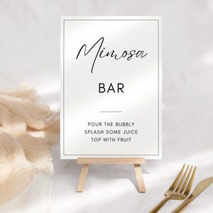May include: White sign with black text that reads "Mimosa Bar" and instructions for making a mimosa: "Pour the bubbly, splash some juice, top with fruit". The sign is on a small wooden easel.