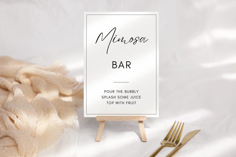 May include: White sign with black text that reads "Mimosa Bar" and instructions for making a mimosa: "Pour the bubbly, splash some juice, top with fruit". The sign is on a small wooden easel.