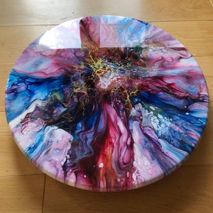 May include: A round, abstract painting with a glossy finish. The artwork features a vibrant mix of colors, including blue, pink, red, and gold, creating a dynamic and fluid design. The piece is displayed on a wooden surface.
