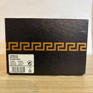Puede incluir: Una caja negra con un dise&ntilde;o de borde geom&eacute;trico dorado. La caja tiene una etiqueta con el nombre de la marca "Versace" y otro texto. La caja est&aacute; sobre una superficie de madera marr&oacute;n claro.