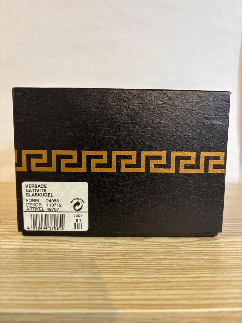 Puede incluir: Una caja negra con un dise&ntilde;o de borde geom&eacute;trico dorado. La caja tiene una etiqueta con el nombre de la marca "Versace" y otro texto. La caja est&aacute; sobre una superficie de madera marr&oacute;n claro.