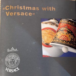 Puede incluir: P&aacute;gina de cat&aacute;logo con el texto "Christmas with Versace" en letras doradas. La p&aacute;gina presenta un logotipo de Versace y una imagen de una campana decorativa con detalles dorados, rojos y negros. La campana tiene un badajo blanco.