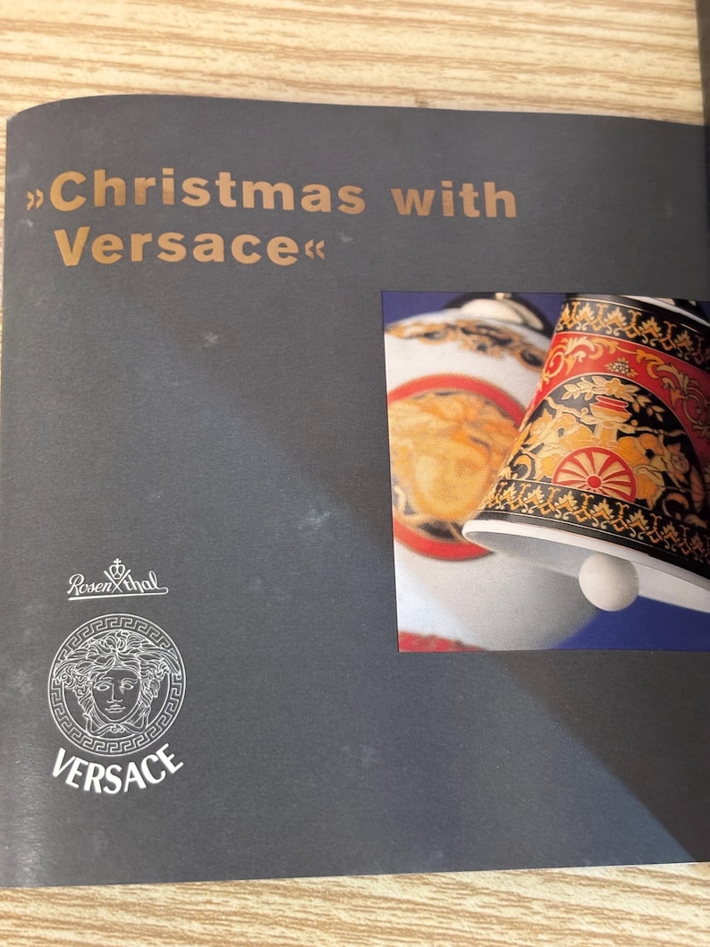 Puede incluir: P&aacute;gina de cat&aacute;logo con el texto "Christmas with Versace" en letras doradas. La p&aacute;gina presenta un logotipo de Versace y una imagen de una campana decorativa con detalles dorados, rojos y negros. La campana tiene un badajo blanco.