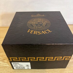 Puede incluir: Una caja negra de Versace con detalles dorados. La parte superior de la caja presenta el logotipo de Versace y la palabra "VERSACE" en letras doradas. La caja tiene una superficie texturizada y un dise&ntilde;o de greca en la parte inferior.