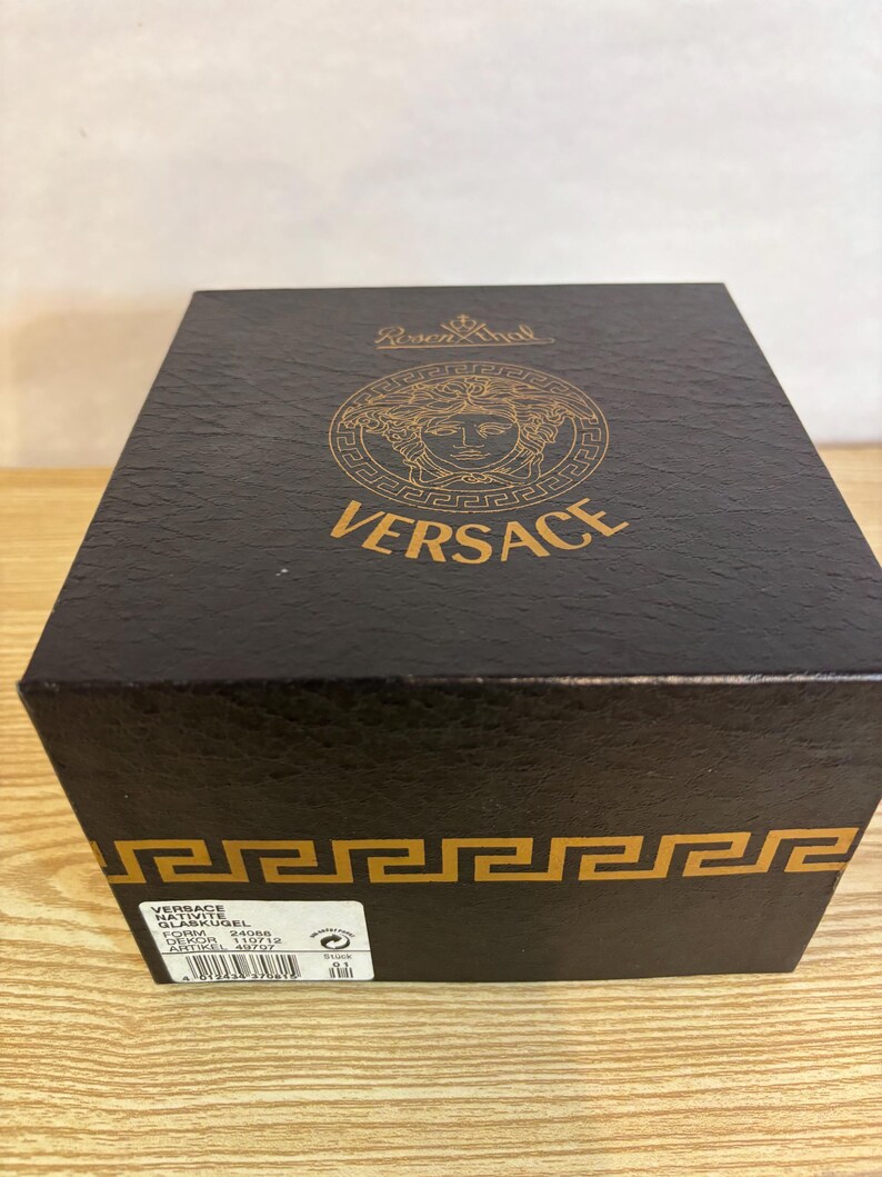 Puede incluir: Una caja negra de Versace con detalles dorados. La parte superior de la caja presenta el logotipo de Versace y la palabra "VERSACE" en letras doradas. La caja tiene una superficie texturizada y un dise&ntilde;o de greca en la parte inferior.