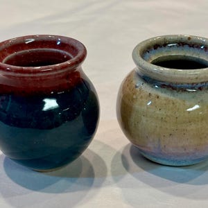 May include: Two small, round ceramic vases. One is glazed in shades of red, dark blue, and teal. The other vase has a tan and blue glaze. Both have a wide opening and a rounded body.