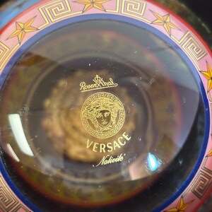 Puede incluir: Un cuenco decorativo de cristal transparente con el logotipo dorado de Versace y el texto "Versace Nativite". El borde presenta un dise&ntilde;o rosa y azul con estrellas doradas y patrones geom&eacute;tricos.