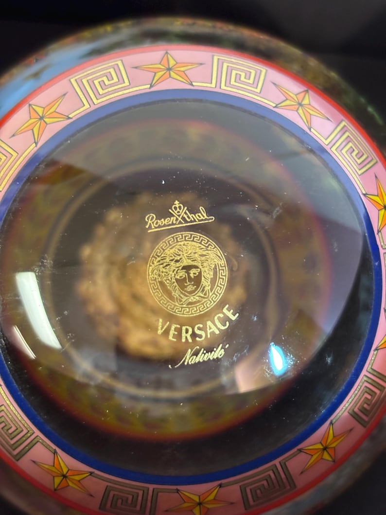Puede incluir: Un cuenco decorativo de cristal transparente con el logotipo dorado de Versace y el texto "Versace Nativite". El borde presenta un dise&ntilde;o rosa y azul con estrellas doradas y patrones geom&eacute;tricos.
