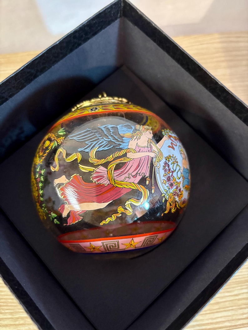Puede incluir: Adorno decorativo y redondo con fondo negro, que presenta una figura alada con atuendo rosa y rojo, sosteniendo un escudo. El adorno est&aacute; alojado en una caja negra, con detalles dorados y un patr&oacute;n de estrellas y geom&eacute;trico.