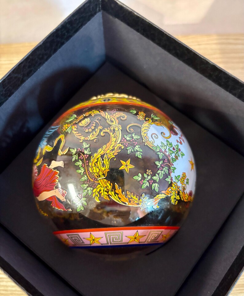 Puede incluir: Una caja de baratijas esf&eacute;rica decorativa con un dise&ntilde;o detallado y colorido. La caja presenta una base negra con detalles dorados, rojos y azules. El dise&ntilde;o incluye motivos florales y de estrellas, y una figura con un vestido rojo.
