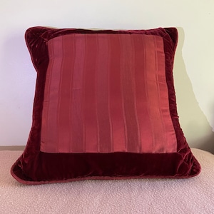Pode incluir: Uma almofada de veludo bordô com um inserto de tecido listrado bordô. A almofada tem uma borda decorativa ao redor da borda.