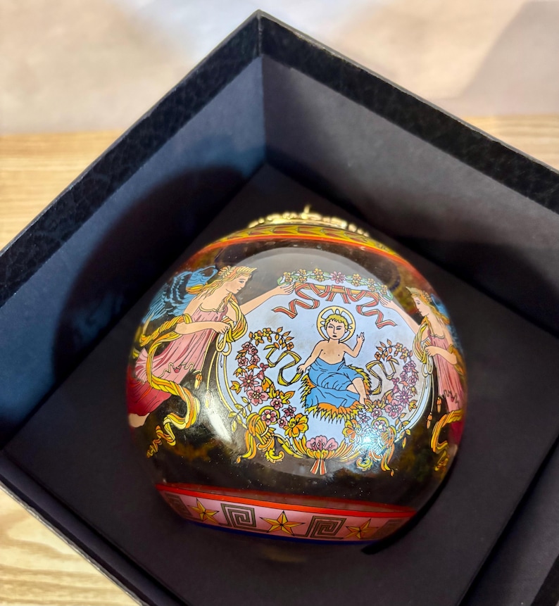 Puede incluir: Adorno redondo decorativo con un dise&ntilde;o pintado detallado. El adorno presenta una imagen central de una figura en azul, rodeada de motivos florales y angelicales en rosa y dorado. El adorno se presenta en una caja negra.